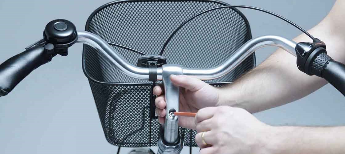 A pair of hands adjust a bicycle stem with an attachment handle in front of a black bicycle basket. The image shows a close-up of the handlebar and the mechanism used to fix or adjust the height and angle of the handlebar.