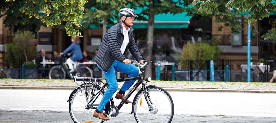 Mand med hjelm og blazer cykler gennem byen – i baggrunden ses caféer, grønne træer og andre cyklister.