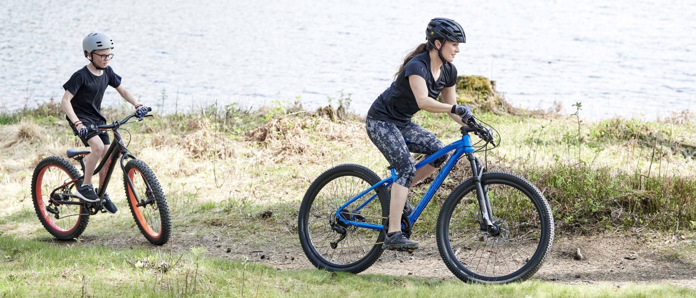 To børn cykler langs en sø på grussti. De er iført cykelhjelme og sportstøj, og omgivelserne er præget af natur og lys sommerstemning.