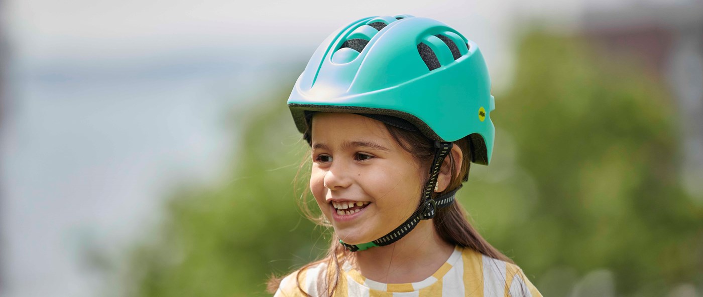 Smiling girl with long hair wearing a turquoise bike helmet. The background is blurred and bright, giving the image a friendly and summery atmosphere. 