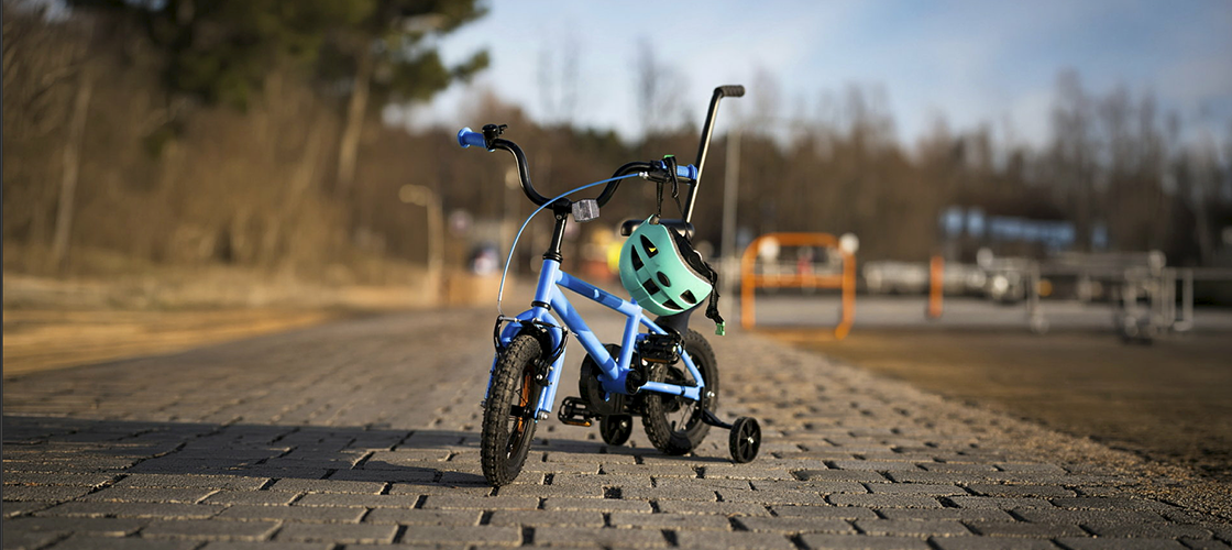 En blå børnecykel med støttehjul står parkeret på en brolagt vej udendørs. En turkis cykelhjelm hænger på styret, hvilket understreger fokus på sikker cykling for børn. Baggrunden er blødt sløret med træer og omgivelser i naturligt dagslys.