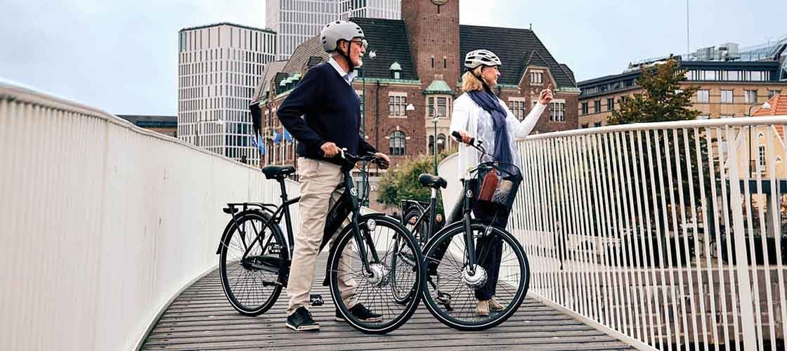 Two adult cyclists take a break on a bridge in the city, with modern and historic buildings in the background, while they chat and enjoy the view.