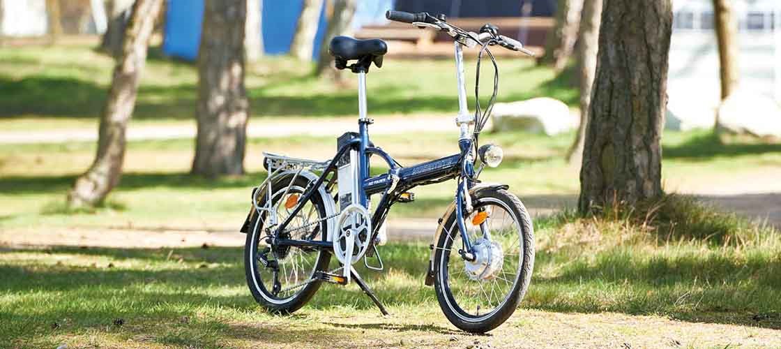 Folding bike parked on grass between trees in a sunny park with buildings in the background.