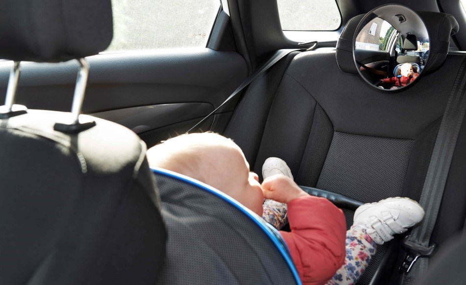 An infant is in a rear-facing car seat in the back seat of a car. The child is wearing red trousers and white shoes. A round rear-view mirror is mounted on the front headrest so the driver can see the child while driving.