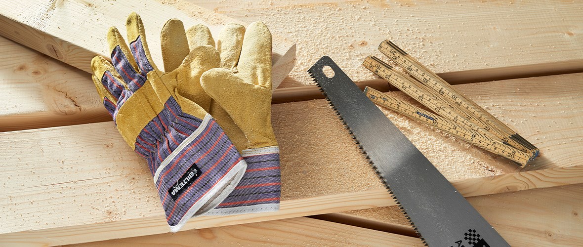 A pair of work or safety gloves, a handsaw and a ruler lie on a stack of light boards covered with sawdust - ready for use in a woodworking project.