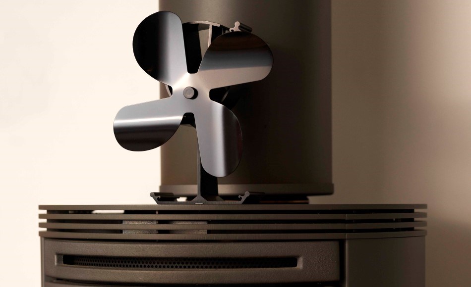 Sort varmemølle til brændeovn med fire ventilatorblade placeret oven på en brændeovn med lamelrist, op ad en lys væg i et minimalistisk interiør.