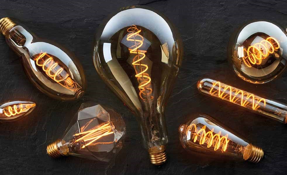 Dekorative glødepærer med synlige filamenter i forskellige former, oplyst og liggende på en mørk overflade.