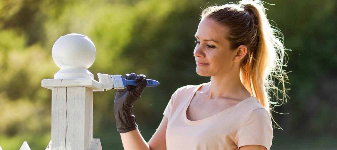 Kvinde i lyserød trøje smiler og mens hun maler en hvid træstolpe i grønne omgivelser.