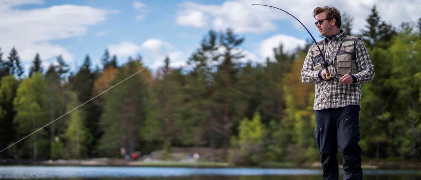 Mand iført ternet skjorte og fiskevest står ved en skovsø og kaster med fluestang. Bag ham ses tæt skov og en blå himmel med spredte skyer.