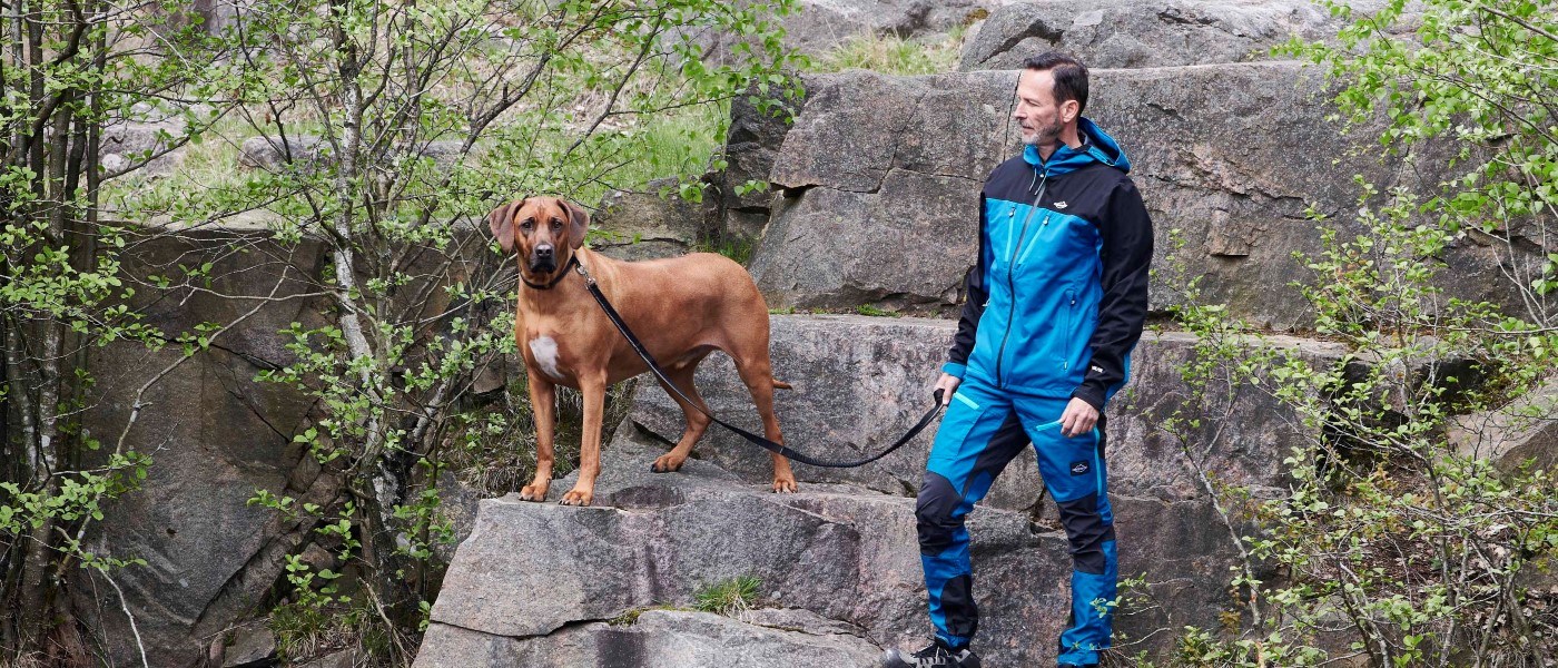 En person i blåt friluftstøj står på klipper i et grønt naturområde og holder en hund i snor. Hunden står på en stenafsats og ser fremad, mens snoren går mellem hunden og personen. Store klippeblokke, buske og træer omgiver området.