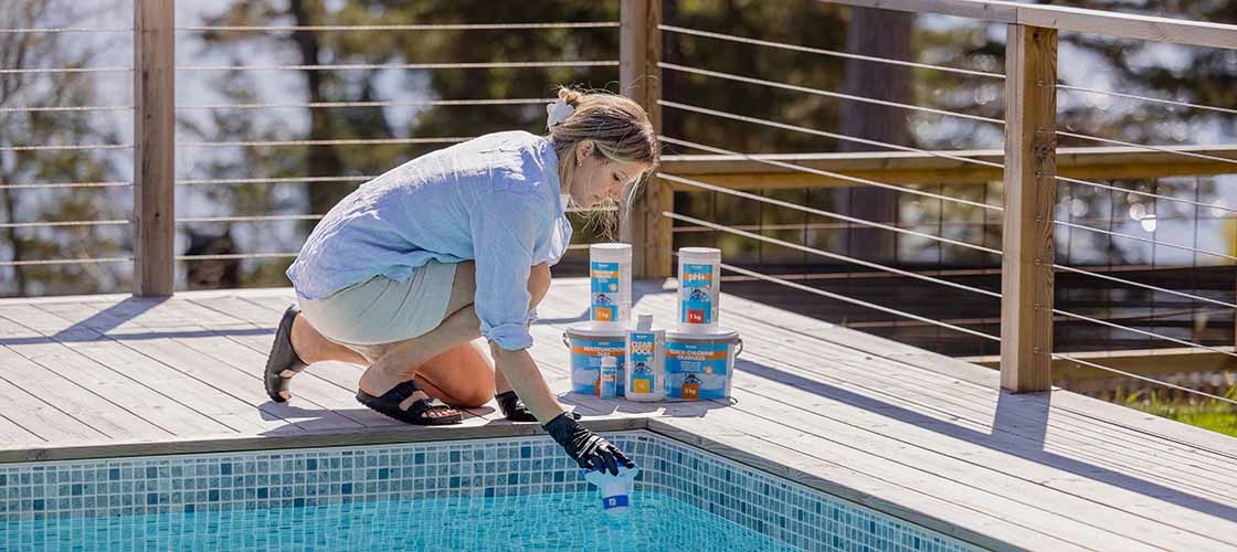 En person iført beskyttelseshandsker tester poolvandet ved kanten af ​​en udendørs swimmingpool. Adskillige beholdere med poolkemikalier står på trædækket ved siden af ​​poolen.