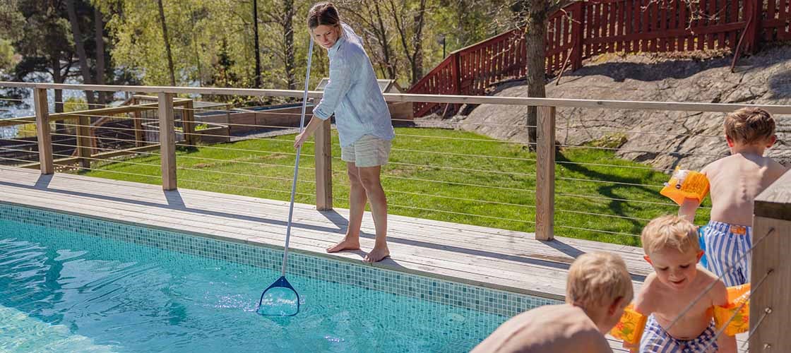 En kvinde står ved kanten af ​​poolen og rengør den med et net. Ved siden af ​​poolen leger tre børn iført badetøj og armvarmere.