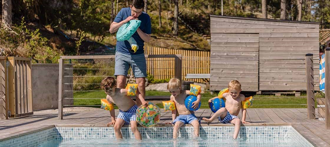 Flere personer sidder og står ved kanten af ​​en udendørs swimmingpool. Tre børn dypper deres fødder i poolen, og en voksen mand står i baggrunden og puster en svømmering op. I baggrunden er en træterrasse, et hegn, et græsareal og en lille bygning omgivet af træer. Billedet viser et sommermiljø, hvor folk er ved en pool for at svømme og slappe af.