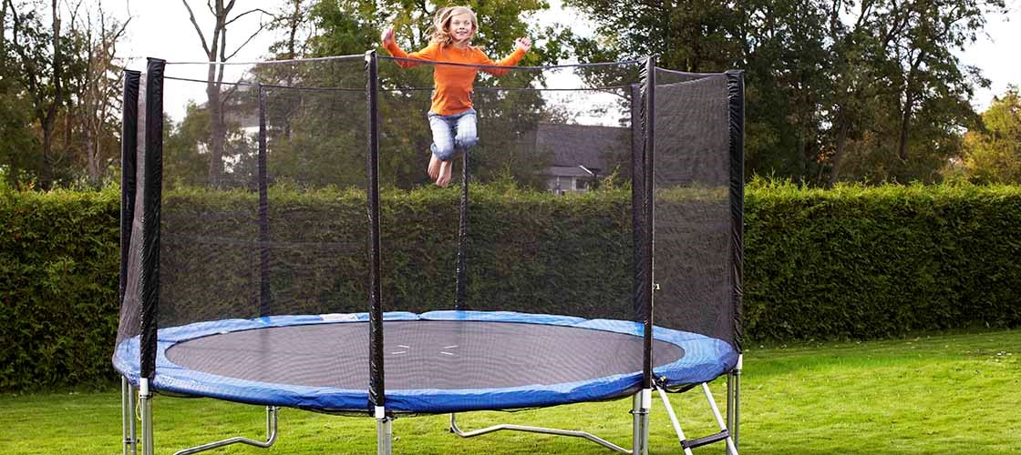 En person hopper på en rund trampolin med et sikkerhedsnet, der er placeret på en græsplæne i en have. Trampolinen har en stige på siden og er omgivet af hække og træer i baggrunden. Billedet viser et udendørsmiljø med fokus på brugen af ​​en trampolin med et sikkerhedsnet for øget sikkerhed.