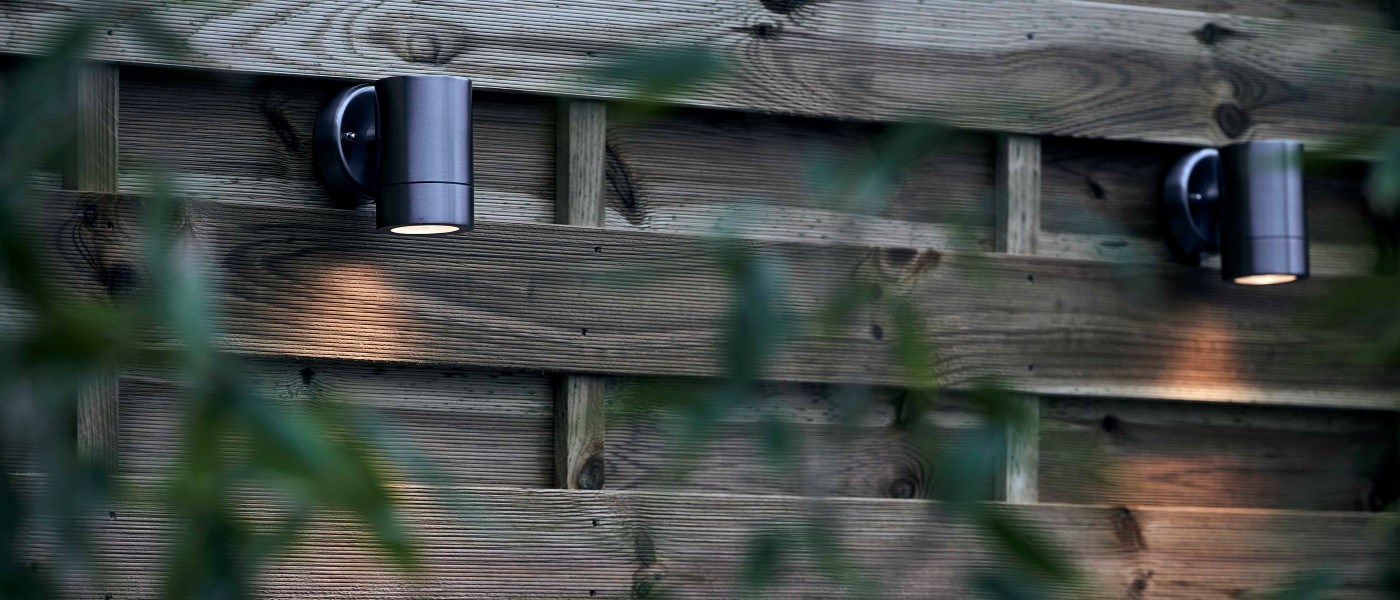 Two modern outdoor lamps from Biltema in black metal are mounted on a vertical wooden slatted wall. The lamps shine downwards and create warm spots of light on the wood, surrounded by green plants in the foreground.