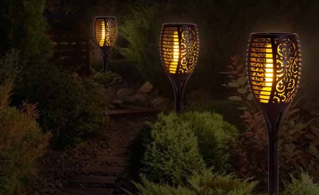 Decorative solar torches light up a garden path in the evening, creating a warm, flickering flame effect among dense green bushes and plants.