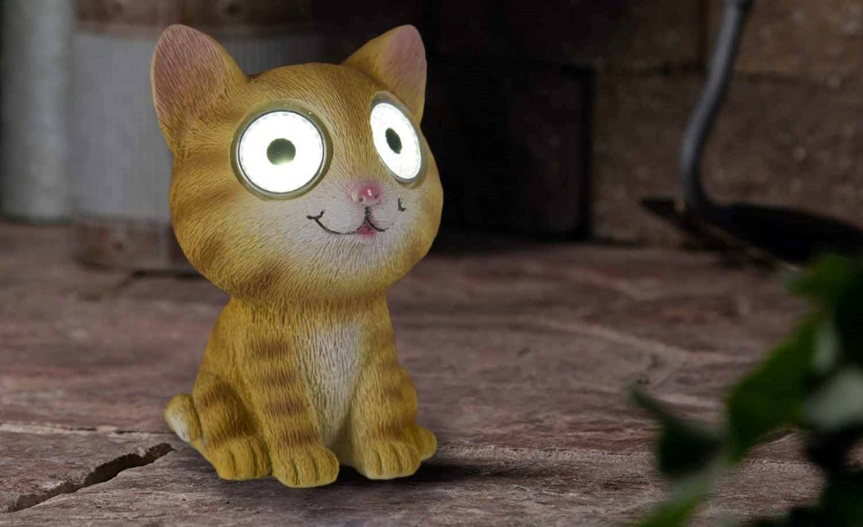 Solar lamp shaped like a cute brown striped cat with big glowing eyes, placed on a tiled terrace next to green plants.