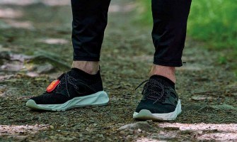 Close-up of a person wearing black running shoes with white soles and black training clothes, walking on a gravel path in green surroundings.