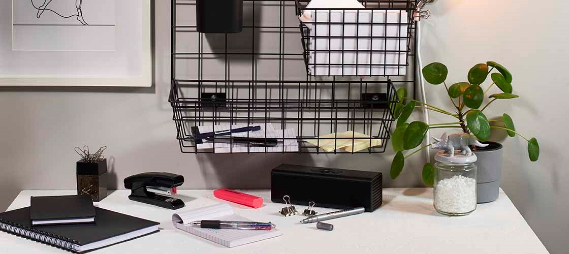 A desk in an office environment with notepads, pens, a stapler, binder clips and a pink highlighter placed on a white work surface. Above the desk is a black wall-mounted metal mesh storage solution with papers, notes and office supplies. On the right, a green potted plant and a glass jar with white contents stand next to a table lamp. The image shows an organized workplace with a focus on storage, order and access to office supplies.