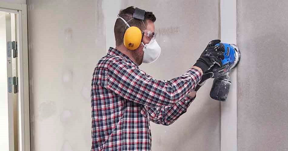 En mand iført ternet arbejdsskjorte, arbejdshandsker, høreværn og ansigtsmaske sliber gipsvægge med en batteridrevet, blå slibemaskine.