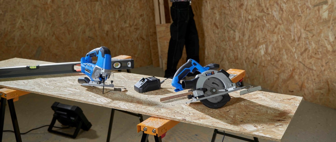 Multi X 18V jigsaw, circular saw and charger from Biltema placed on a workbench in a wood workshop.