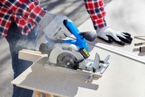 Person wearing a checked shirt and work gloves is using a circular saw with blue handles to cut a wooden board outdoors.