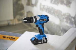 Blue and black Biltema cordless drill stands on a white surface in the middle of a renovation environment with brick walls in the background.