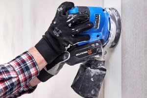 Person wearing a checked shirt and black work gloves is using a blue random orbital sander to sand a wall.