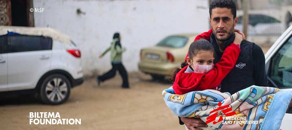 Man holds child wearing winter clothes in relief area with MSF equipment in the background.