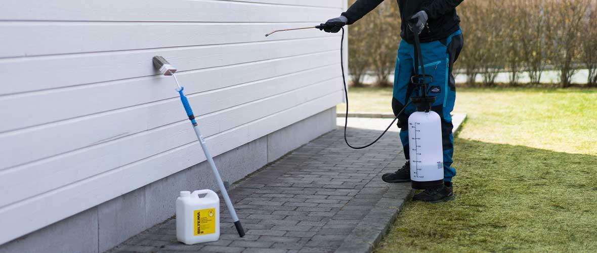 Person spraying facade with wood protection using pressure sprayer, with yellow canister and extension handle standing on tiling on white house wall.