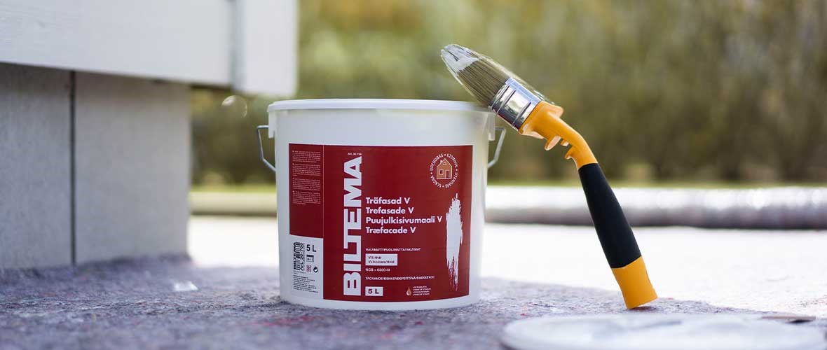 Paintbrush resting on red bucket of outdoor wood protection placed at the base of the house on a sunny day.