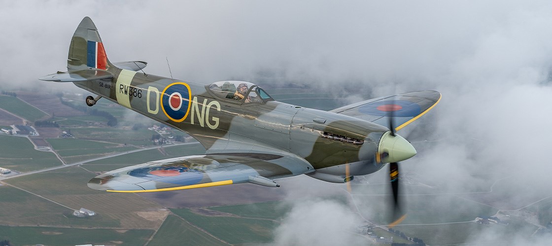 Historisk militærfly af typen Supermarine Spitfire i RAF-farver flyver gennem skyer over landskab.