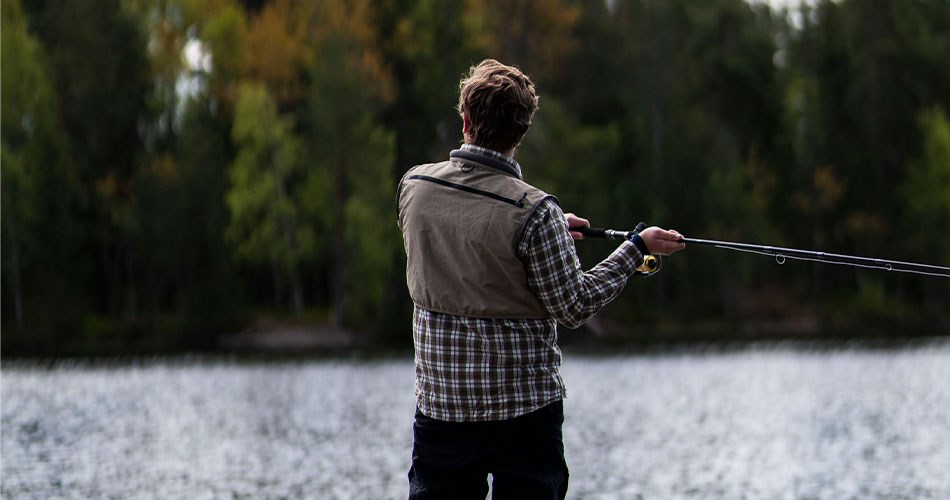 Person standing with his back turned, fishing in a lake surrounded by dense forest and autumn-coloured trees.