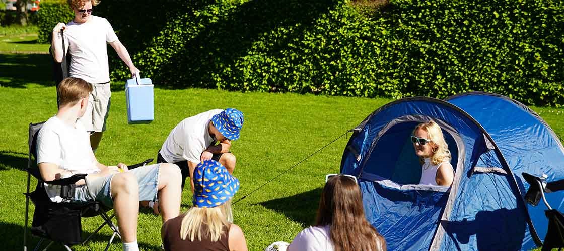 En gruppe personer samles omkring et blåt telt på en græsplæne med køleboks og campingudstyr i solskinsvejr.
