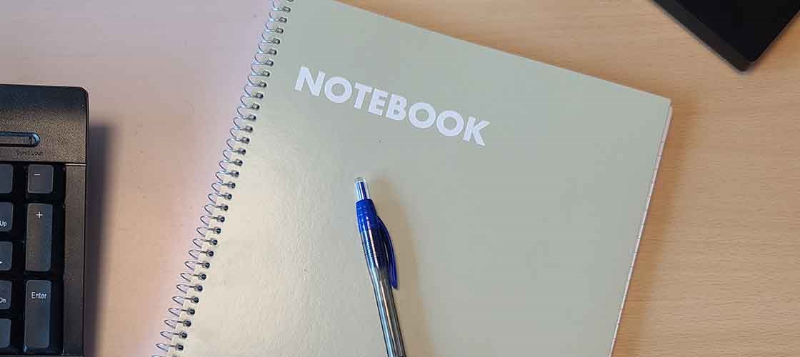 Spiralnotesbog med teksten “NOTEBOOK” og blå kuglepen på skrivebord.
