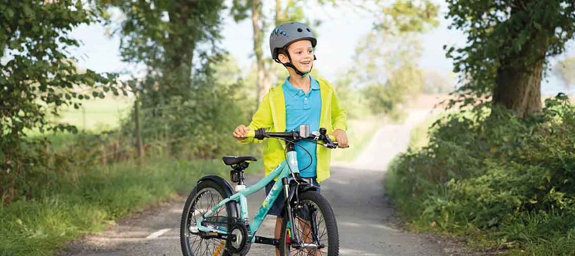 Smilende barn iført cykelhjelm står med sin cykel på en solbeskinnet landevej omgivet af grønne træer og natur.