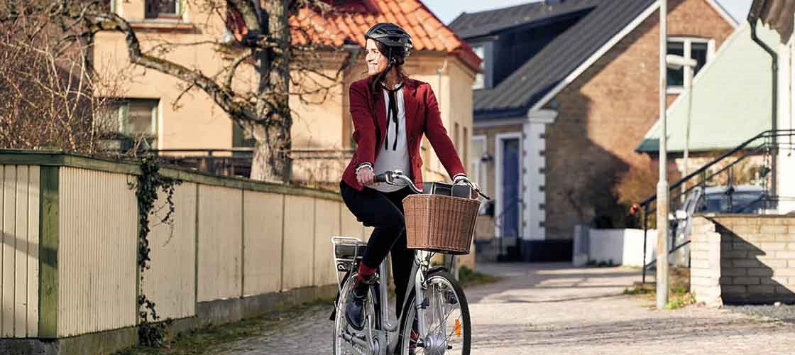 Kvinde i rød jakke cykler gennem et solbeskinnet boligområde med traditionelle huse i baggrunden – hun har cykelhjelm på og en kurv foran på cyklen.