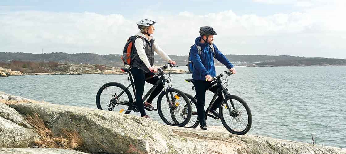 To cyklister med hjelme holder pause på en klippe ved havet og nyder udsigten over vandet og det bakkede landskab i baggrunden.