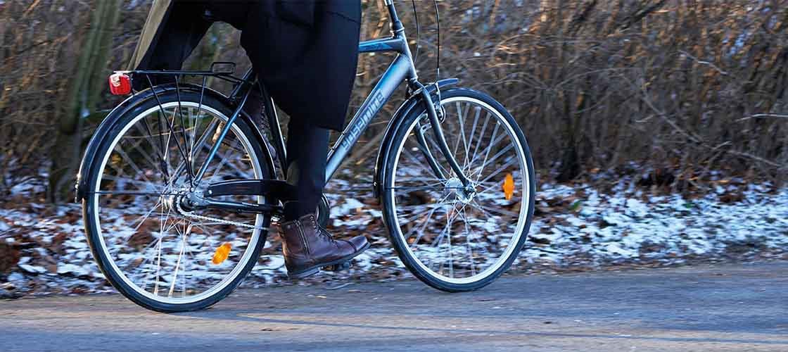 Person cykler på en grå cykel langs en vej med sne og blade langs kanten. Cyklisten bærer mørkt tøj og brune sko, og billedet fokuserer på cyklens hjul og benene i bevægelse.