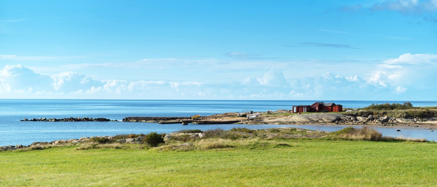 Udsigt over en grøn græsplæne foran et kystlandskab med blå himmel og hav i baggrunden. Der ligger røde hytter eller bygninger tæt ved vandet, og klipperne strækker sig ud i havet.