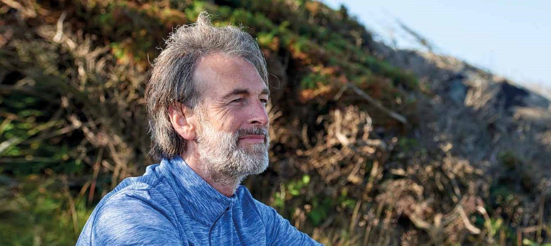 Mand med gråt hår og skæg i blå skjorte sidder udendørs i naturen og nyder udsigten – afslappet og eftertænksom stemning.