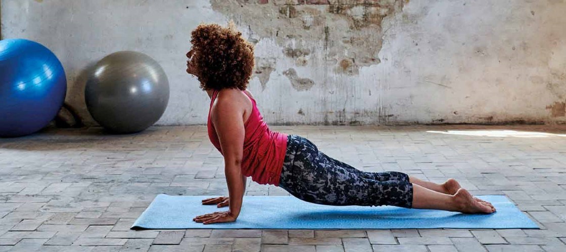 Kvinde udfører yogaøvelsen "opadgående hund" på en blå måtte i et lyst rum med rå vægge og en træningsbold i baggrunden.