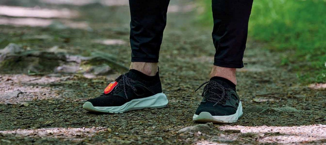 Nærbillede af en person iført sorte løbesko med hvid sål og sort træningstøj, gående på en grussti i grønne omgivelser.